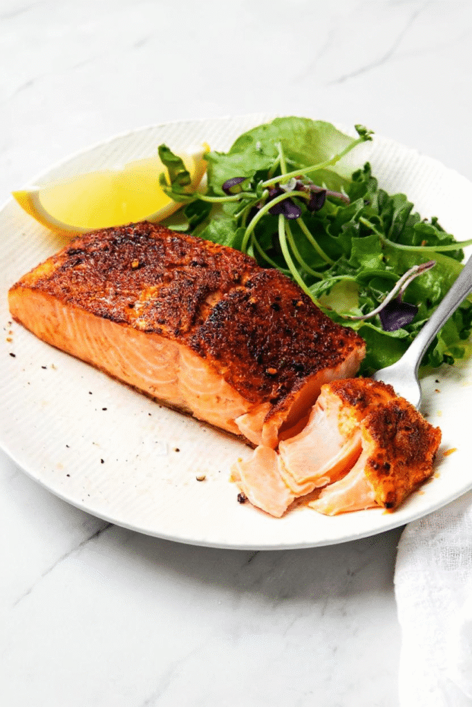 Perfectly cooked air fryer salmon served with greens and lemon on a white plate.