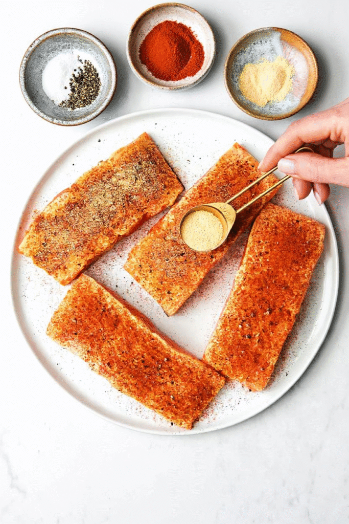Four salmon fillets being seasoned with a dry rub blend including paprika and garlic powder.
