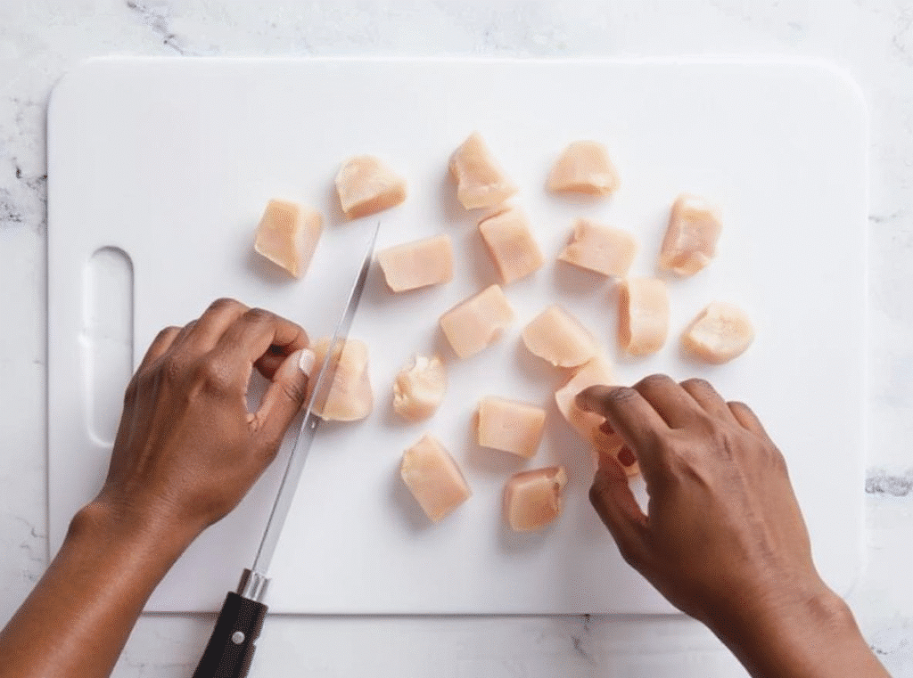 Hands cutting raw chicken breast into bite-sized cubes on a white cutting board.