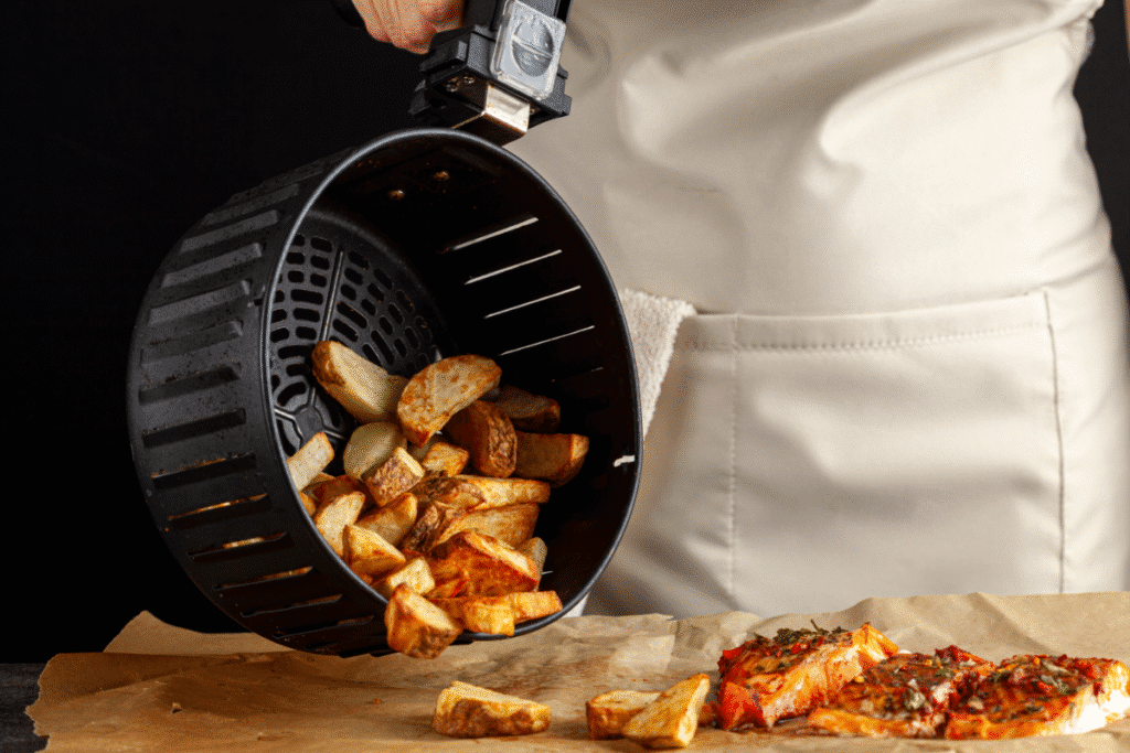 Air fryer basket pouring roasted potato wedges onto parchment beside cooked salmon