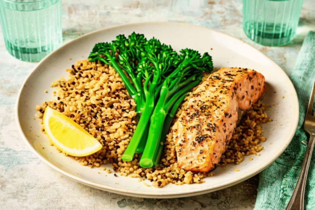 Air fryer salmon fillet served with quinoa and broccolini on a white plate with lemon wedge.